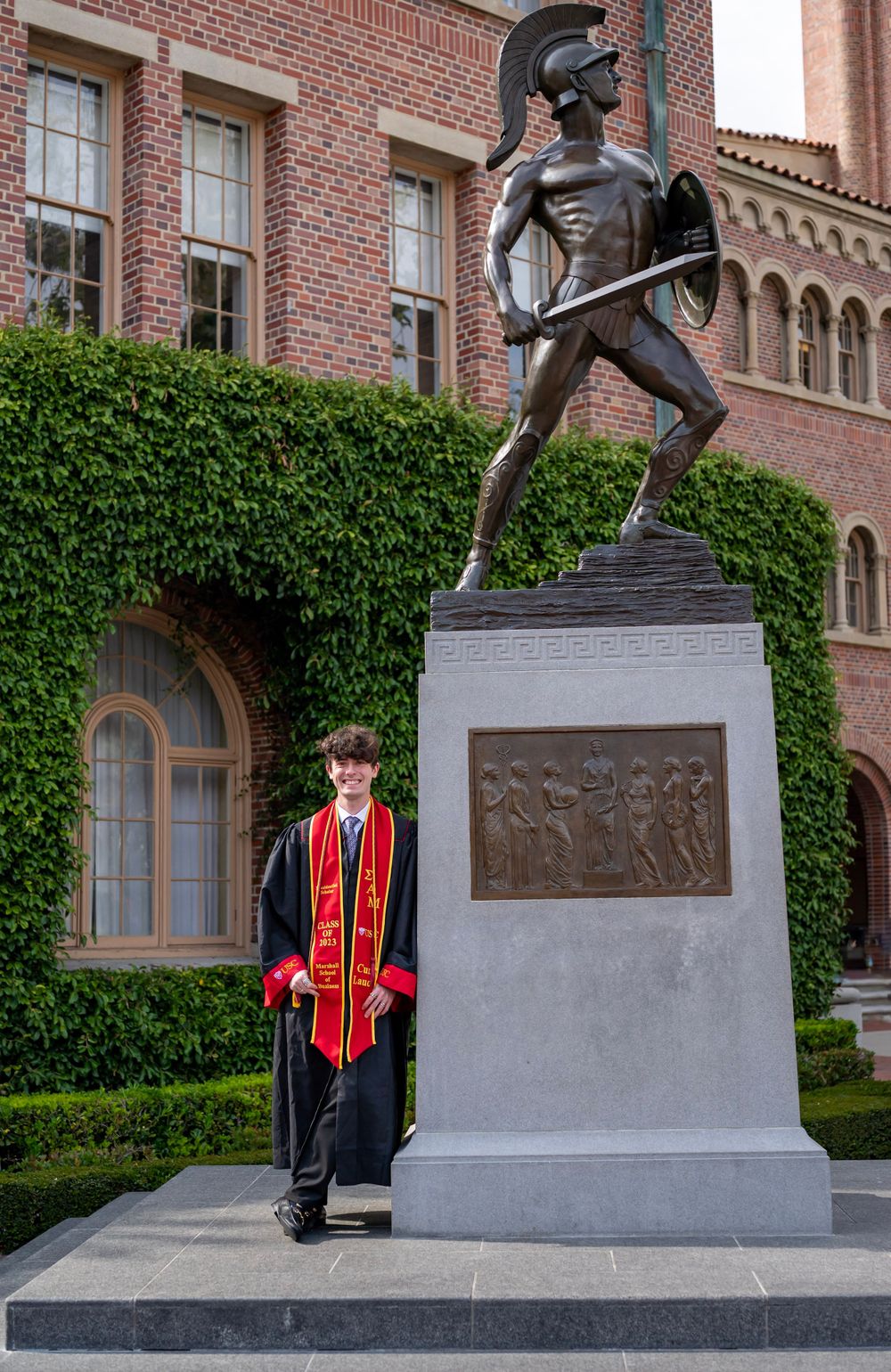 Graduate stands beside a Trojan warrior statue in front of a brick university building.