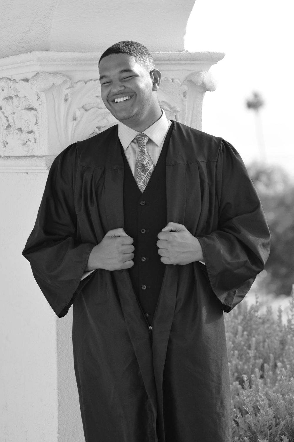 Graduate wearing a black graduation gown and vest smiles warmly in front of a white pillar.