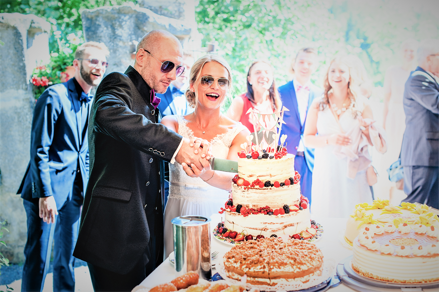 Brautpaar schneidet Hochzeitstorte an - beide haben eine Sonnenbrille auf - Gäste sehen zu