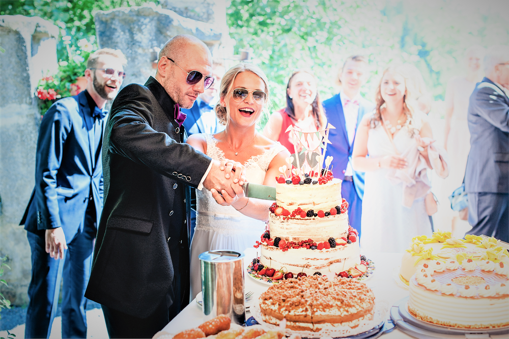 Brautpaar schneidet Hochzeitstorte an - beide haben eine Sonnenbrille auf - Gäste sehen zu