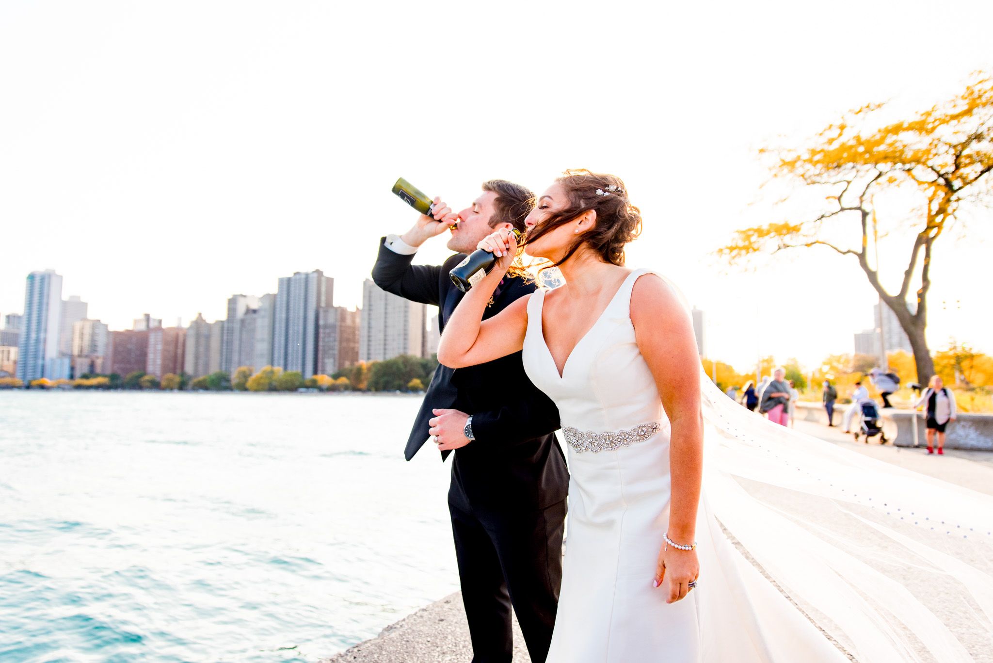bride and groom chug champagne in front of park and chicago skyline at North Avenue beach