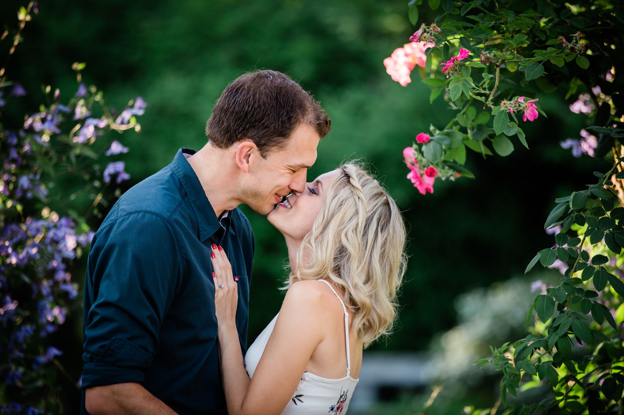 A couple stands in a garden surrounded by purple and pink flowers. They nearly kiss, but stop to smile instead