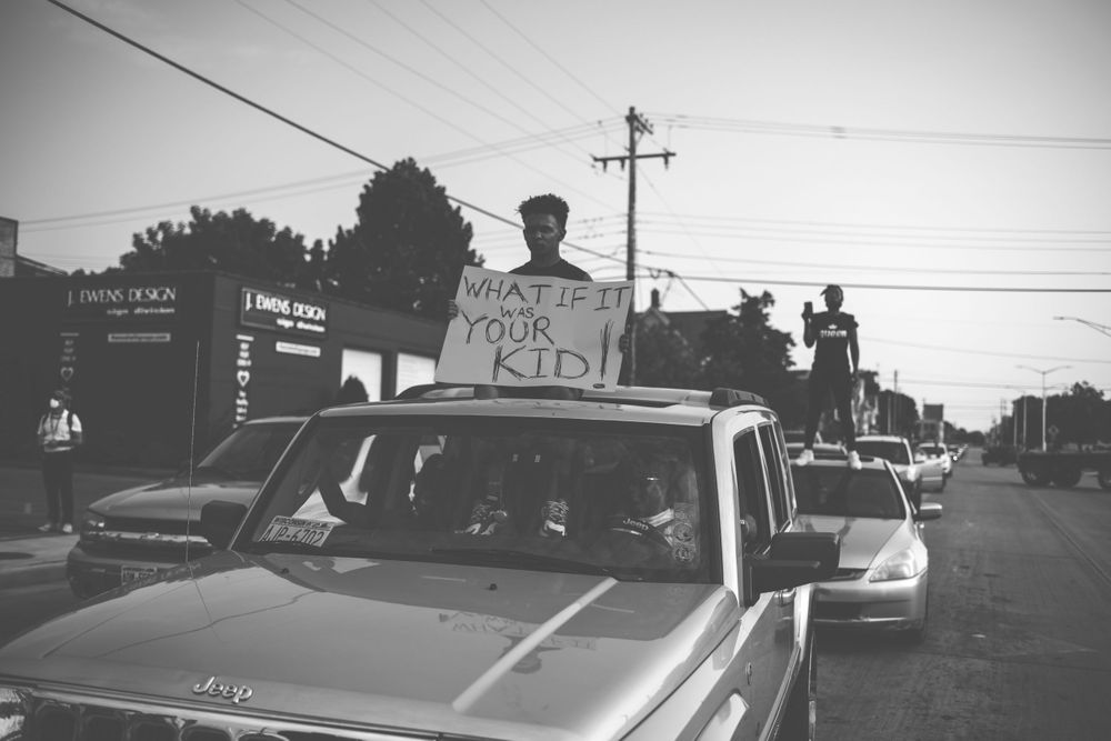A man sits out of the sunroof of a car holding a sign that reads, "What if it was your kid!"