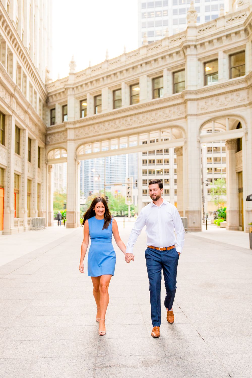 woman with brown hair in a light blue dress holding hands with fiance in front of the wrigley building in chicago