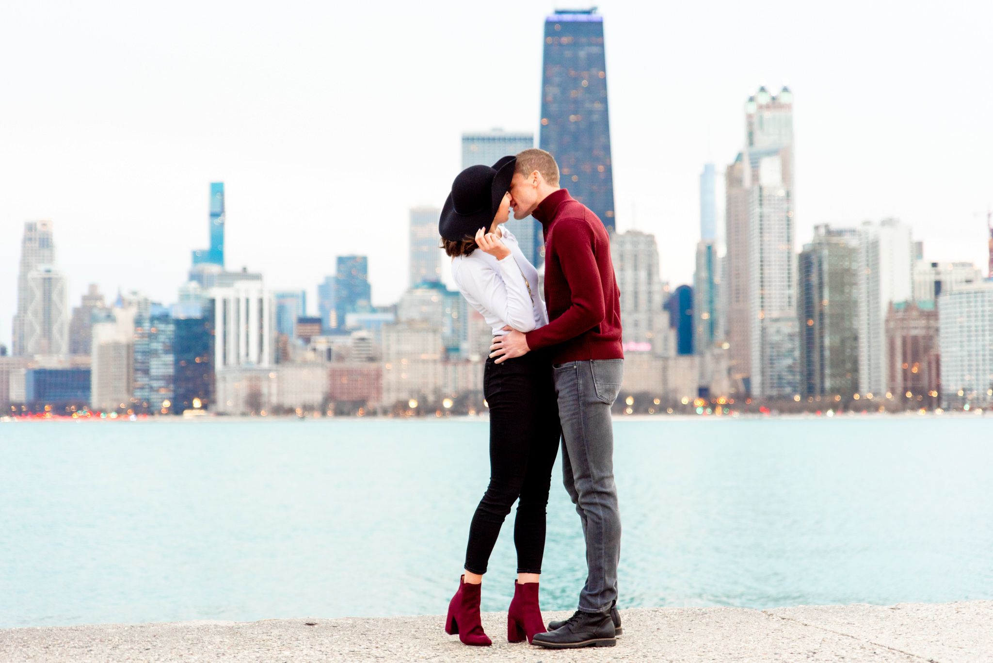 man in read sweater kissing woman in white shirt and black jeans at sunset for Chicago engagement pictures