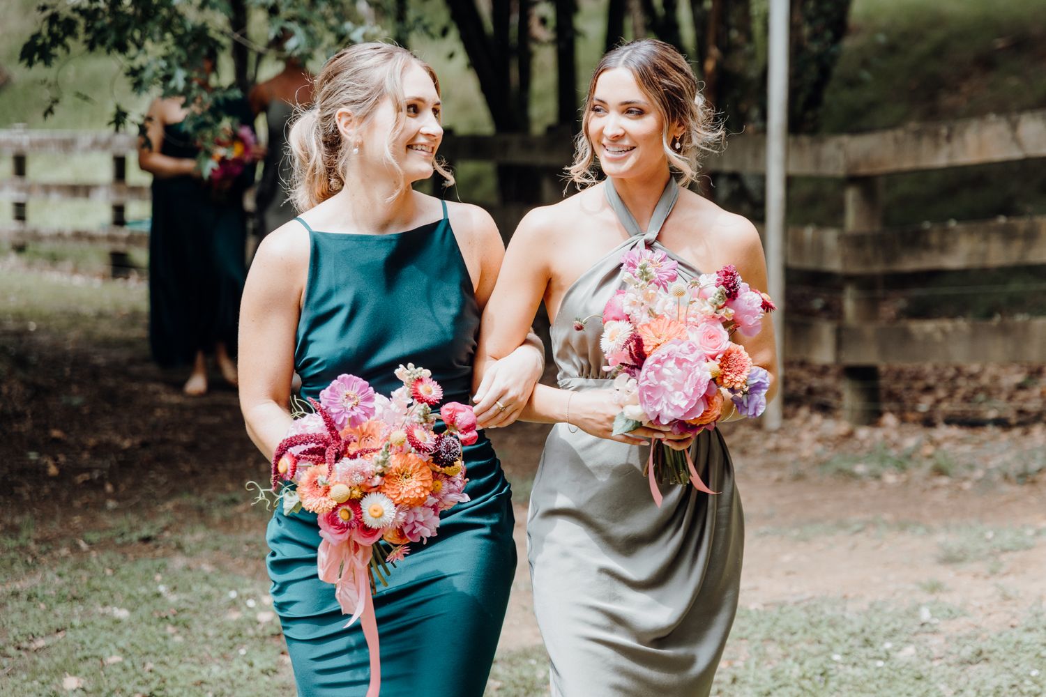 Two attendants in elegant dresses hold pink and coral flower bouquets while walking together.