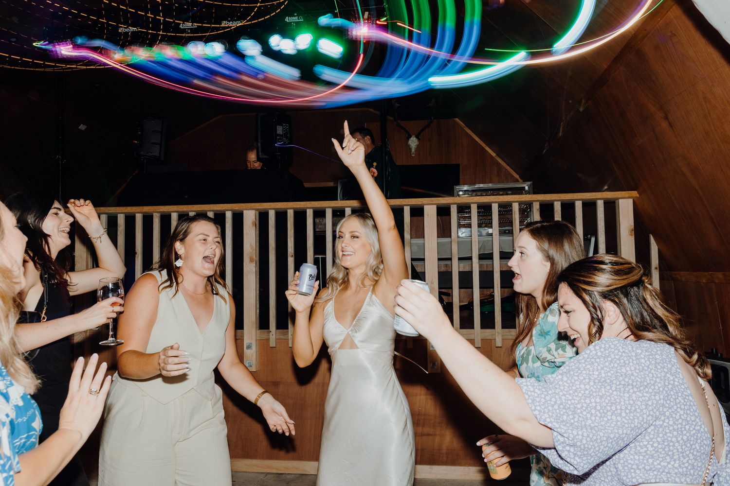 Wedding party dances under colorful neon lights at a rustic indoor venue during reception celebration.
