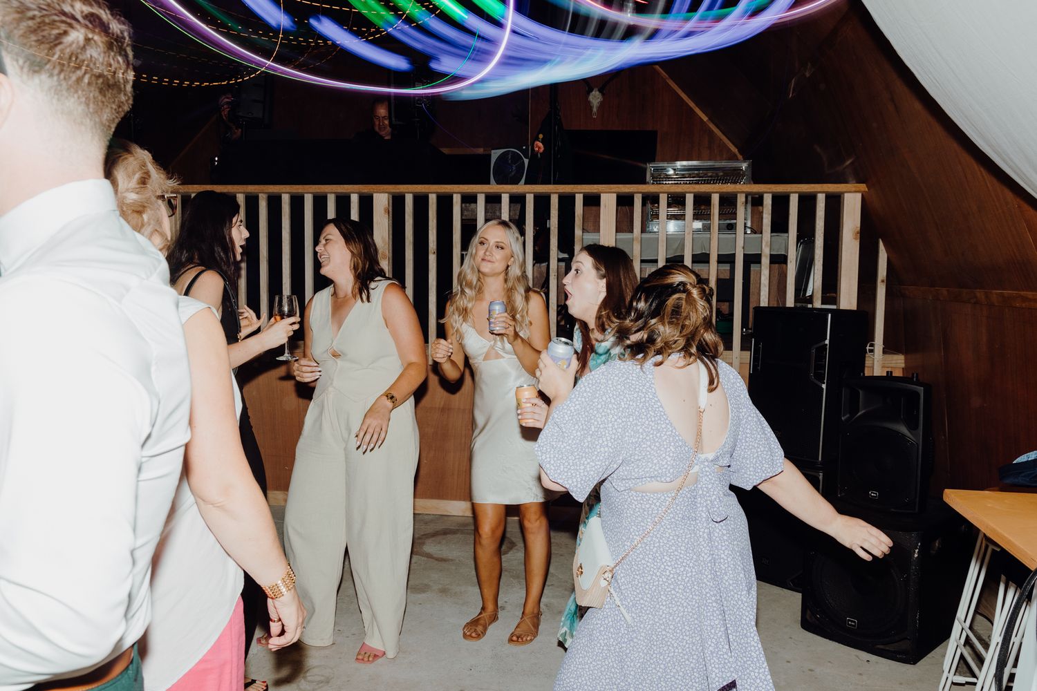 Reception guests dance beneath vibrant LED light trails in a wooden-beamed venue space.