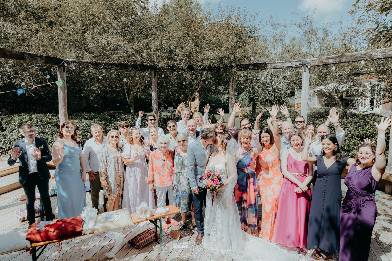 A large wedding party group photo with guests in colorful attire gathered on a wooden deck outdoors.
