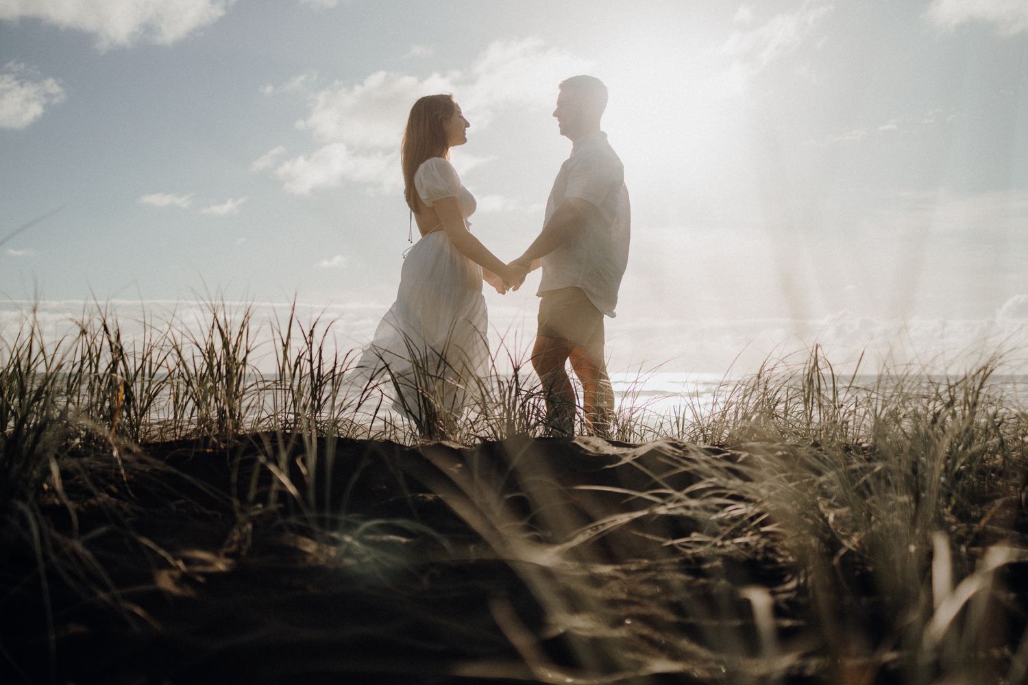 Silhouetted couple holds hands while standing in tall grass at sunset.