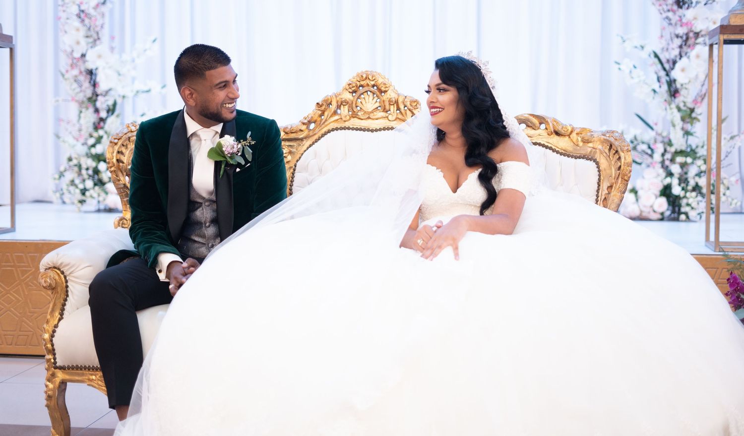 Elegant wedding couple sitting on an ornate gold throne decorated with floral arrangements.