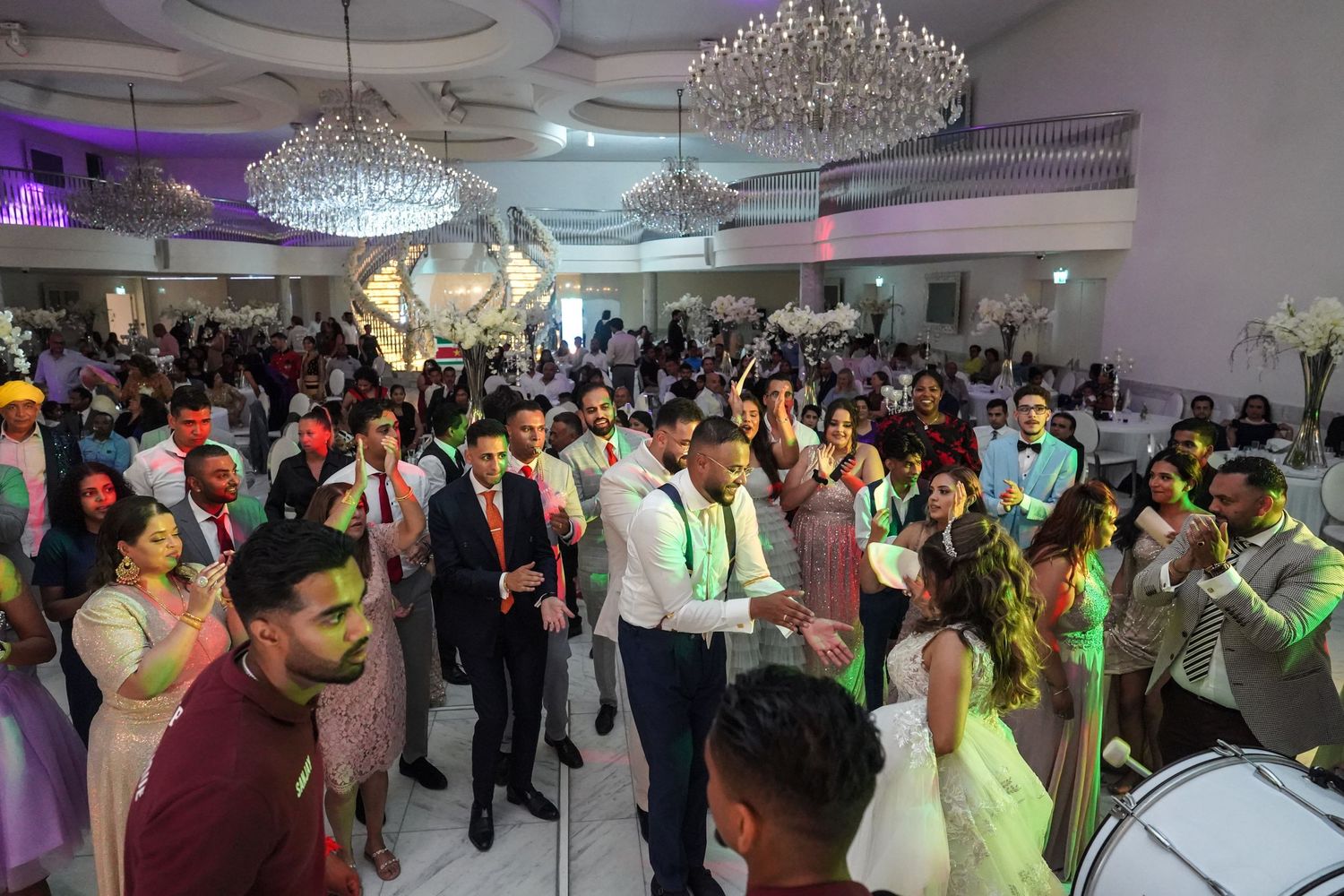 A crowded wedding reception celebration with vibrant lighting and multiple crystal chandeliers overhead.