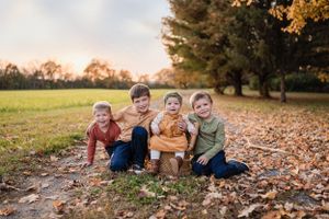 Four children sit together smiling among autumn leaves in park setting.