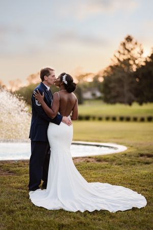 A romantic wedding couple embraces at sunset on a grassy lawn with a trailing white wedding dress.