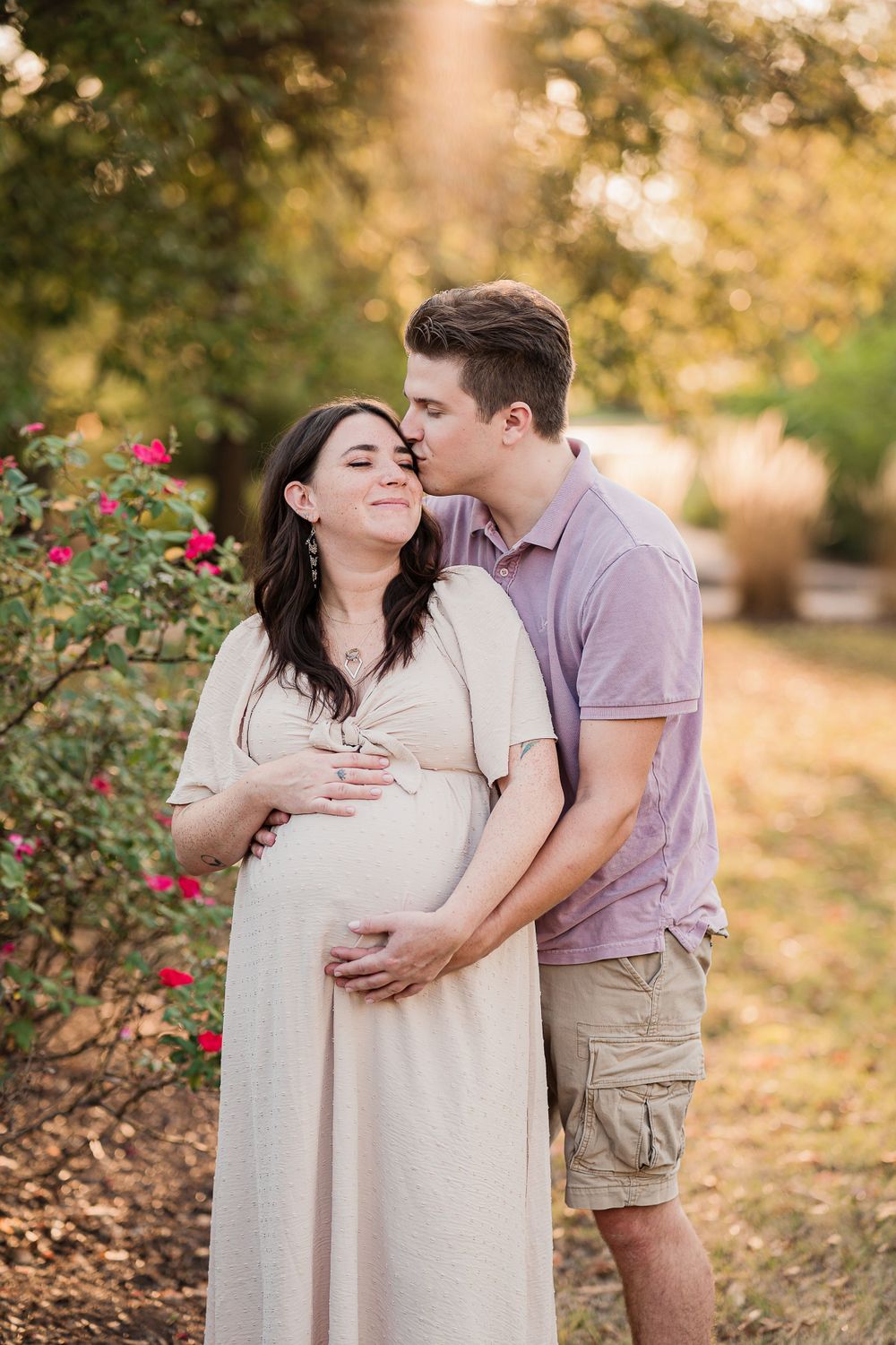 Expecting couple shares tender moment by blooming rose bush in warm evening light.