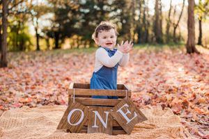 Toddler in blue overalls claps excitedly next to wooden blocks spelling ONE during autumn photo session.