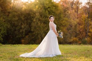 Bride in lace wedding gown poses in a golden autumn field at sunset holding a bouquet.
