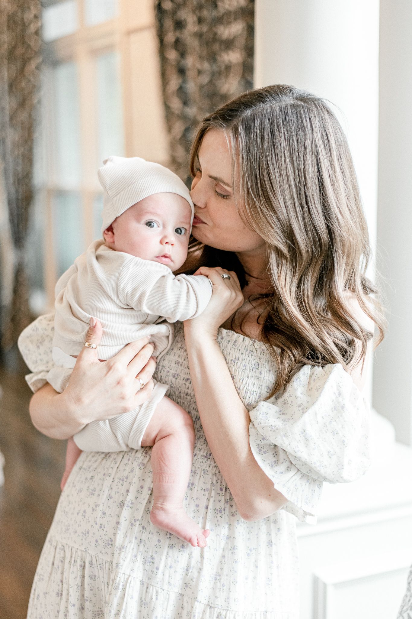 Mother kisses her baby's cheek, baby wears all white with a bonnet on