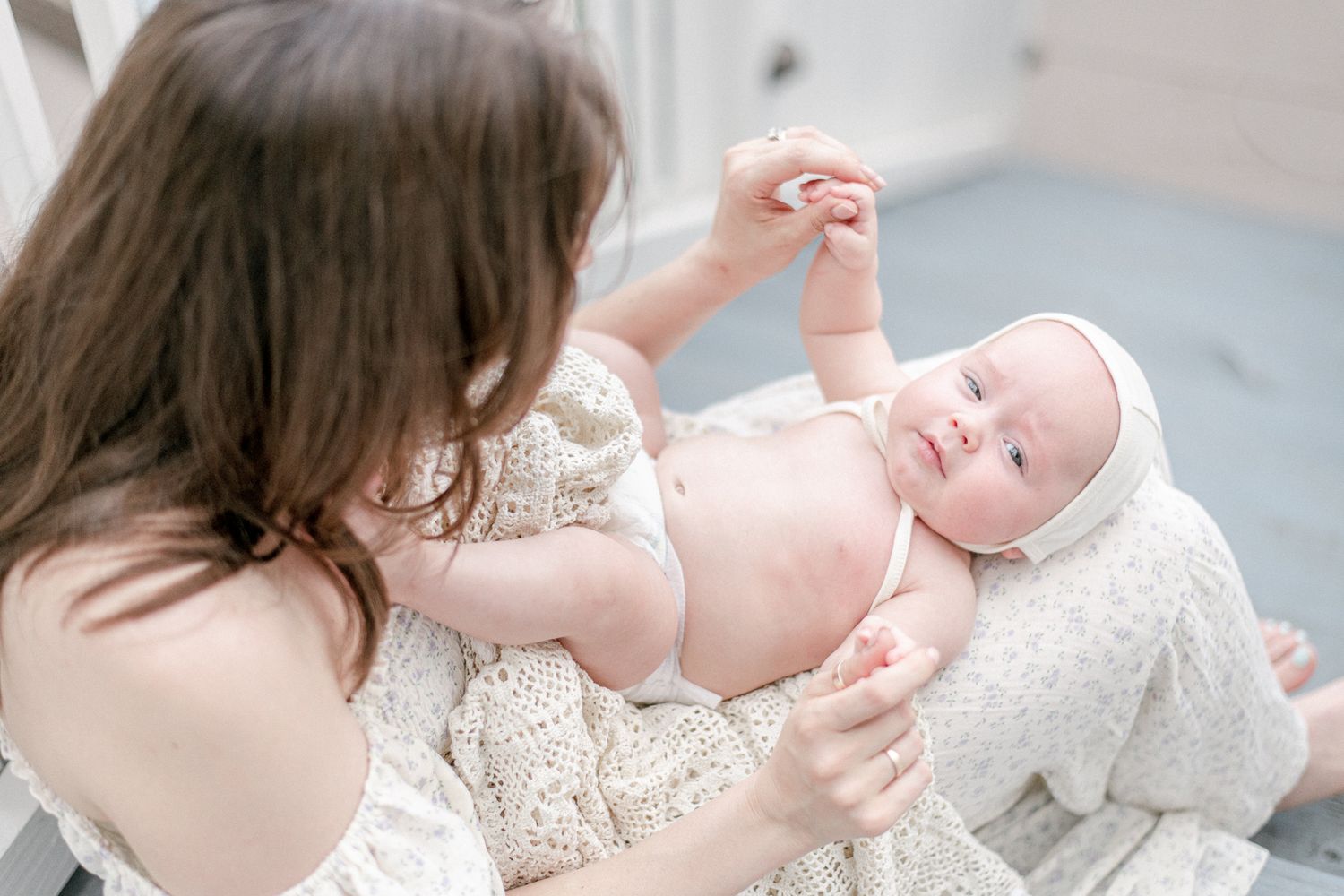 Baby in a bonnet holds his mothers hands as he lays in her lap on the porch