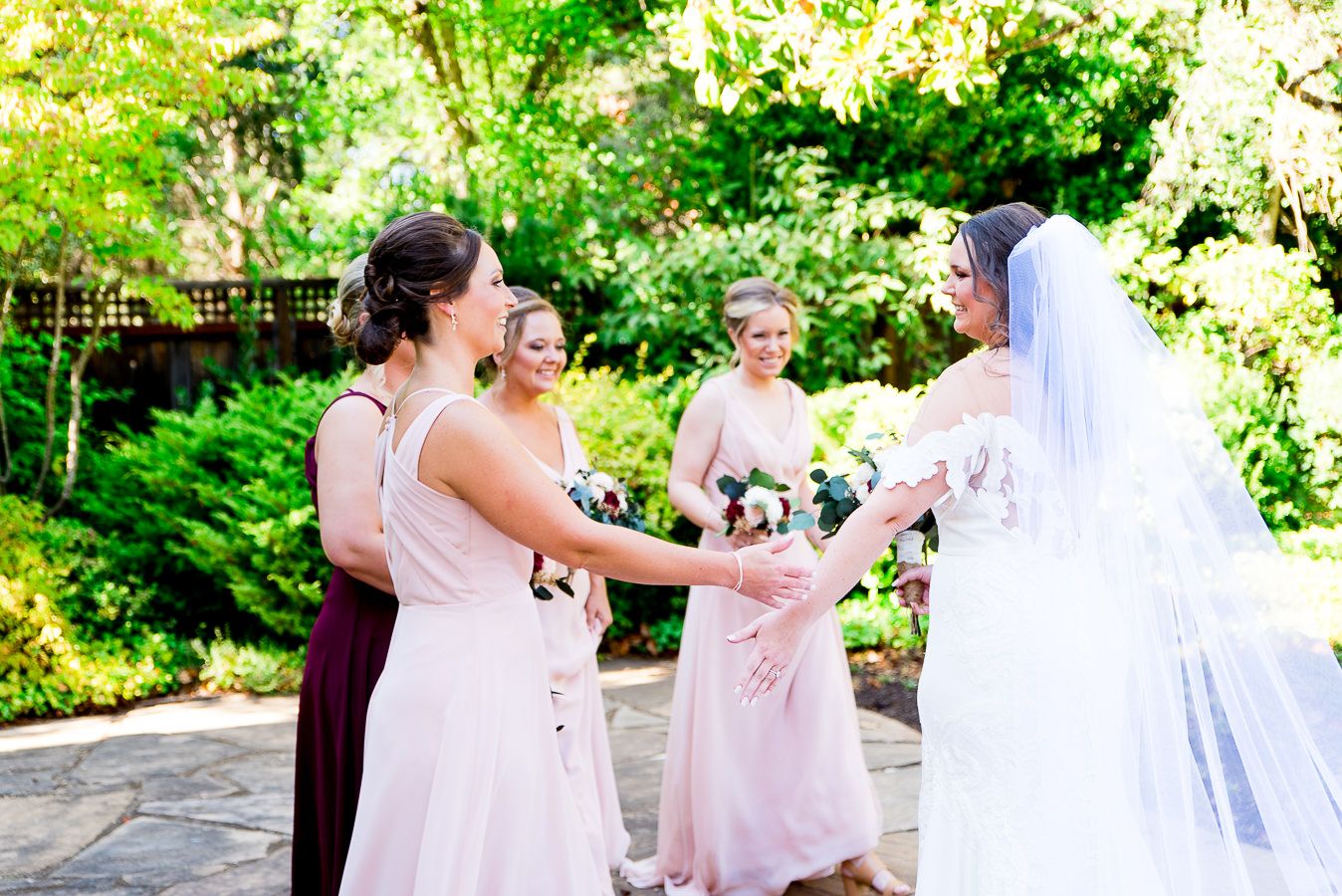 bridesmaids in blush and red hug the bride after they see her for the first time on wedding day