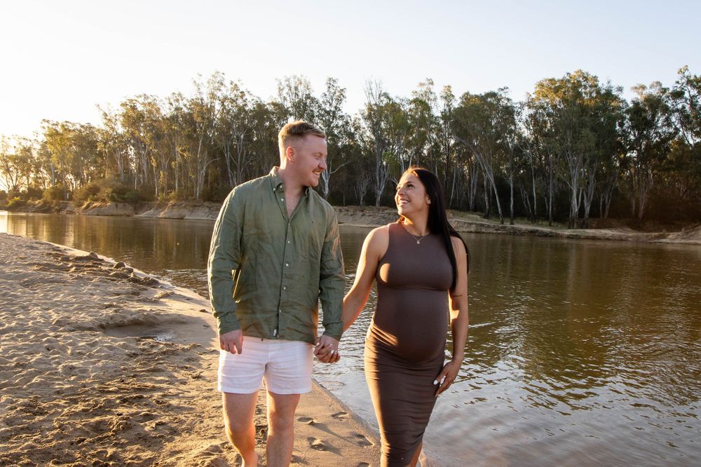 A couple takes a romantic sunset walk along a riverbank with gum trees in the background.