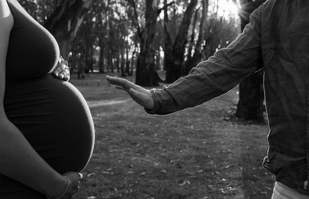 Black and white artistic photograph showing a pregnant belly with a reaching hand in a wooded outdoor setting.