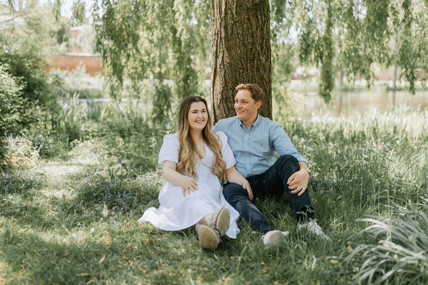 A couple sits under a weeping willow tree on a bright summer day surrounded by tall grass and natural scenery.