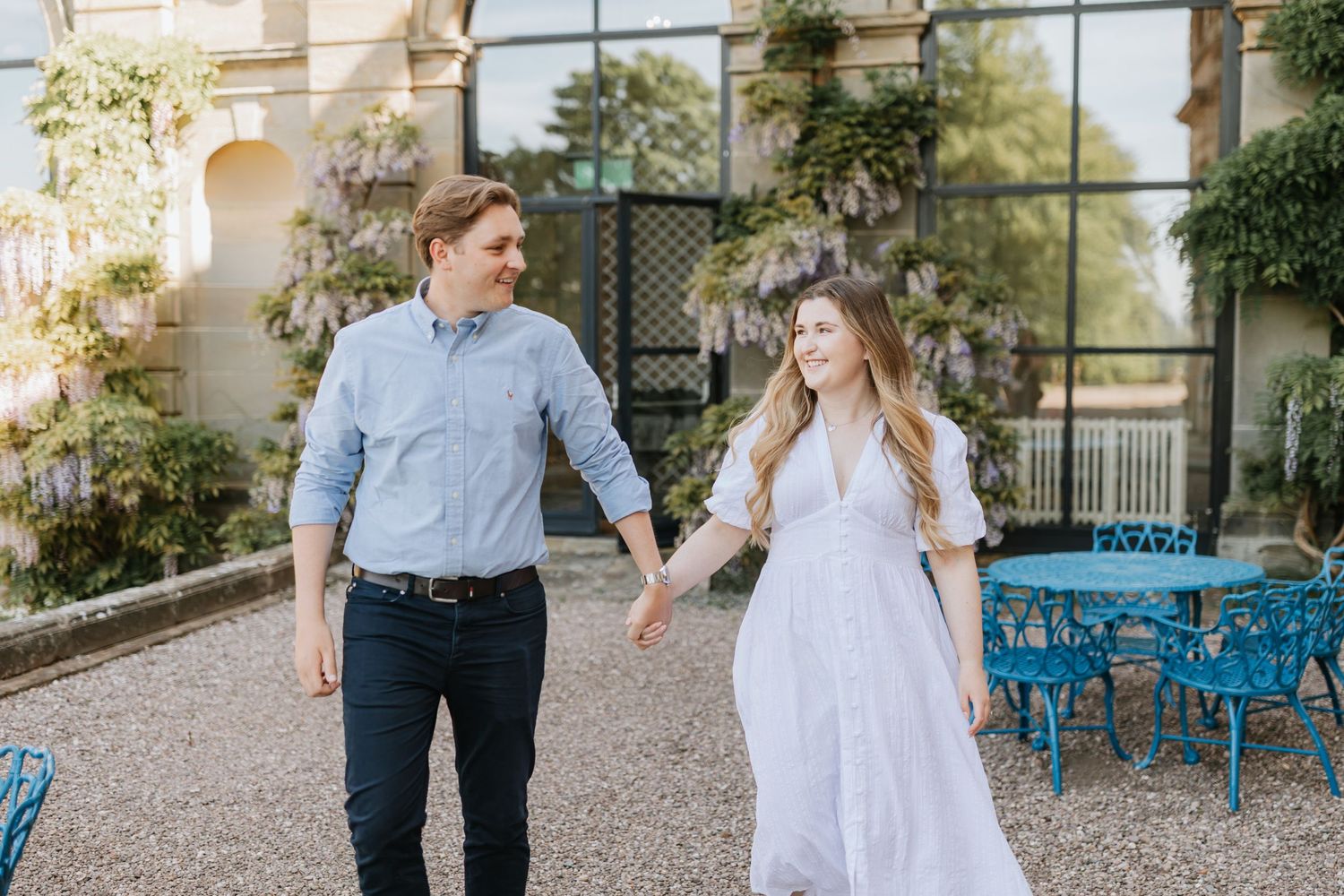 A couple walking hand in hand on a garden patio with bright blue chairs and climbing vines on the wall behind them.