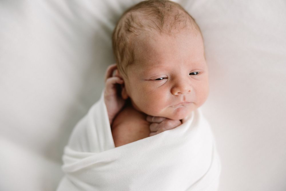 A peaceful newborn baby wrapped in white swaddling cloth against a light background.
