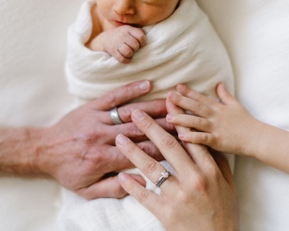 Wedding rings visible as parents' hands gently hold swaddled newborn.