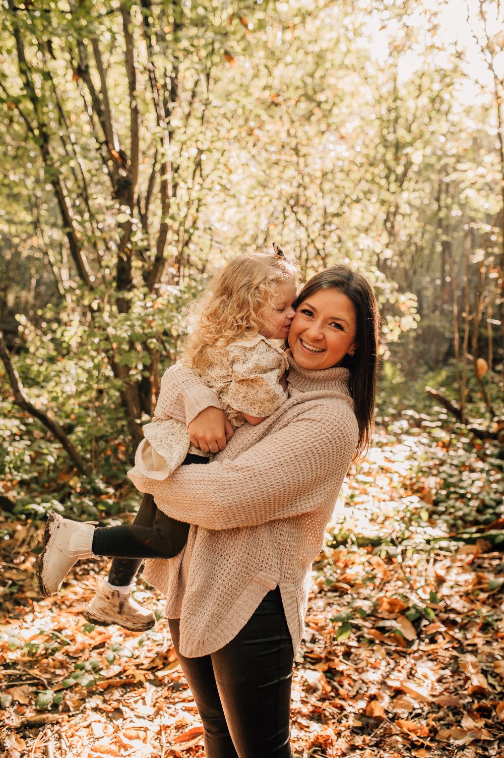 Two people share a joyful embrace while standing among autumn leaves and blooming trees in a sunlit forest setting.