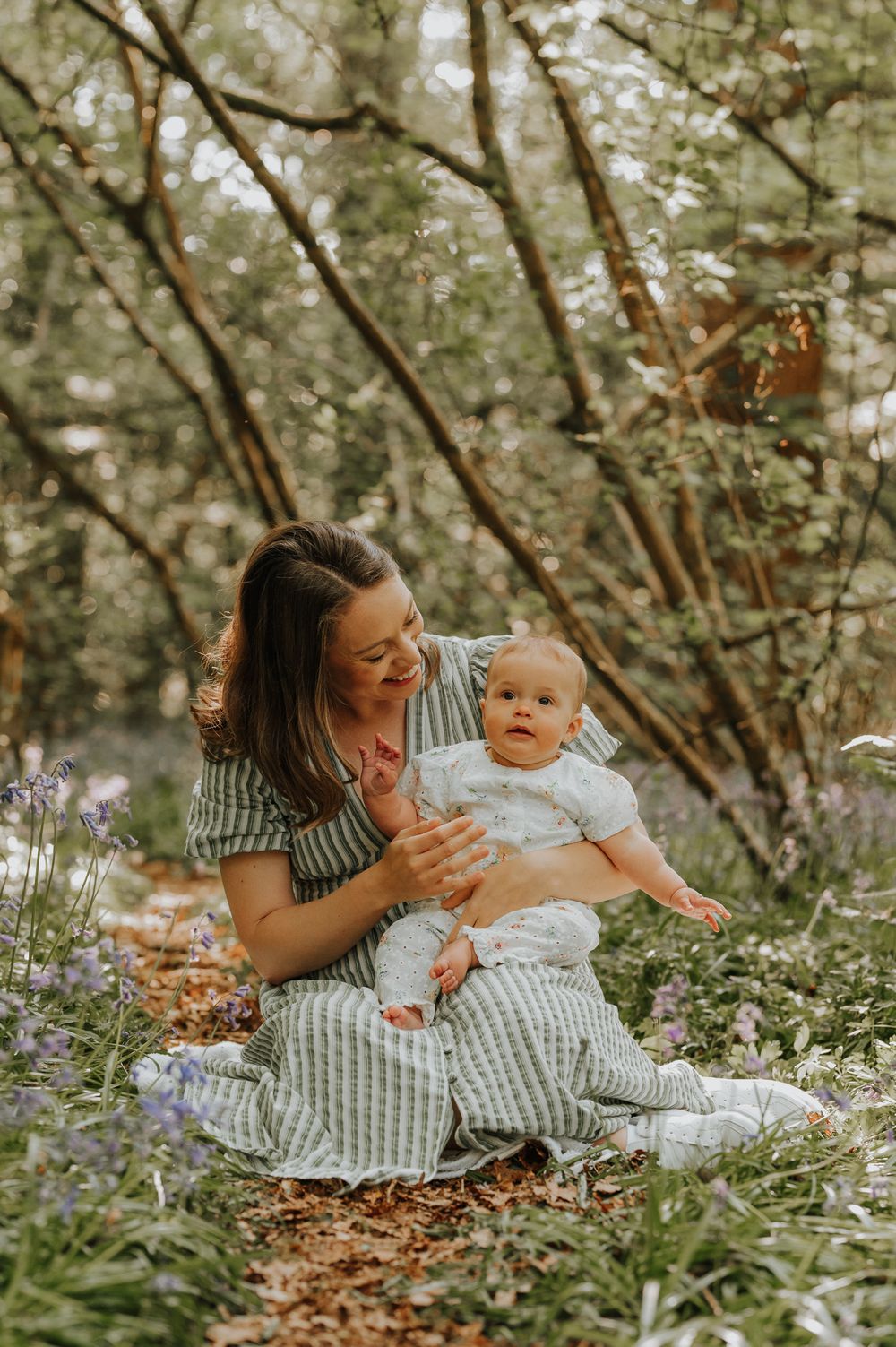 Mother and baby sit together in a spring garden surrounded by blooming purple wildflowers and dappled sunlight.