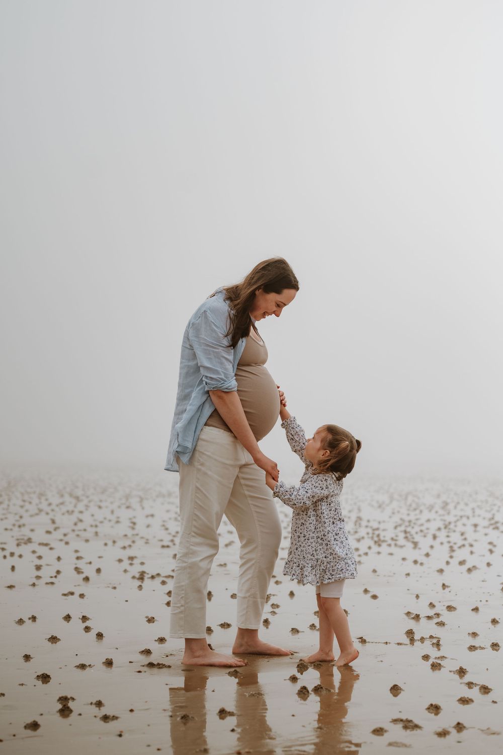 Two people walking barefoot together on a misty beach strewn with seaweed, creating a tranquil coastal scene.