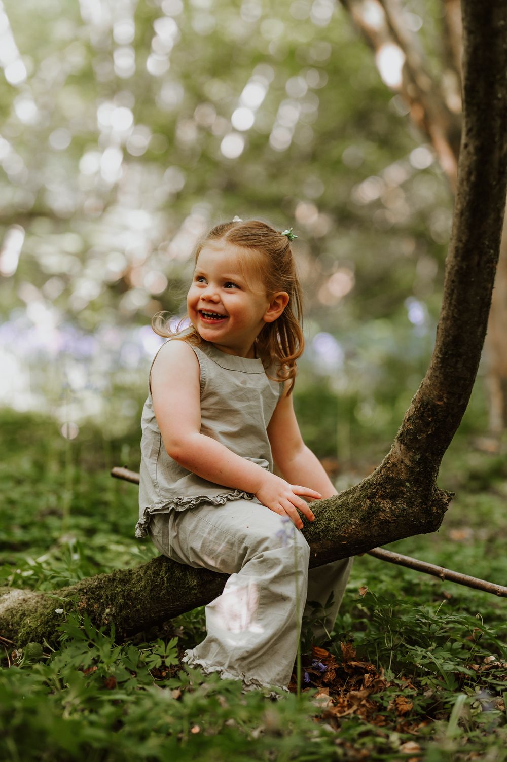 Small child wearing beige outfit sits happily on tree branch in forest with bokeh lighting and green background.