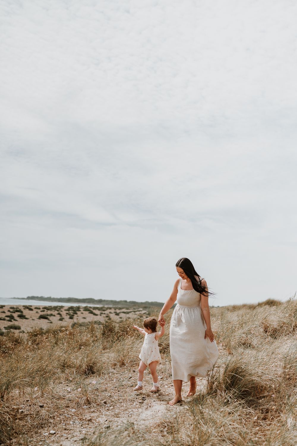 A figure in a white dress holds hands with a small child while walking along a grassy coastal path on an overcast day.