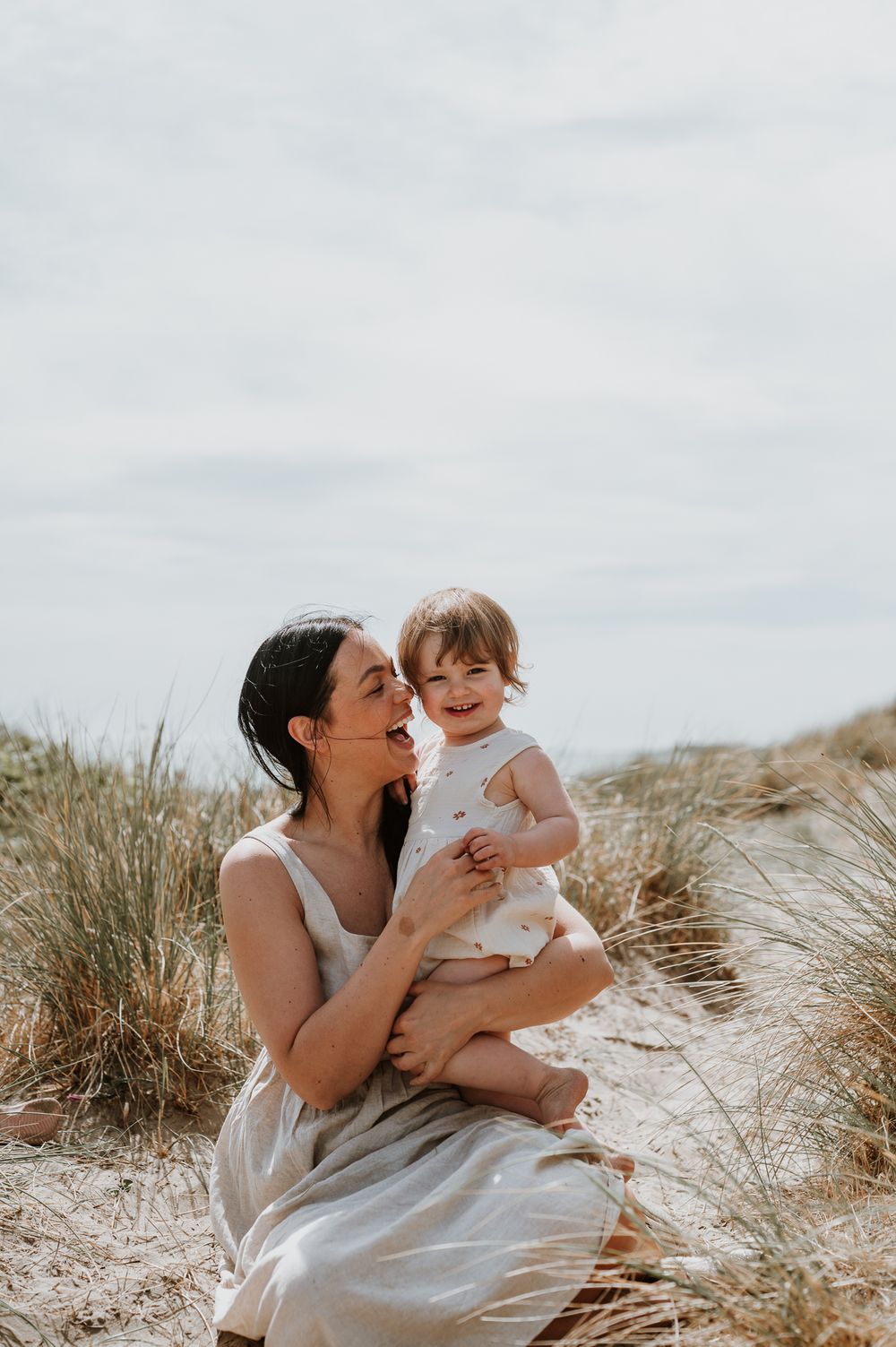 Mother and child share joyful moment together on sandy beach surrounded by dune grass on overcast day.
