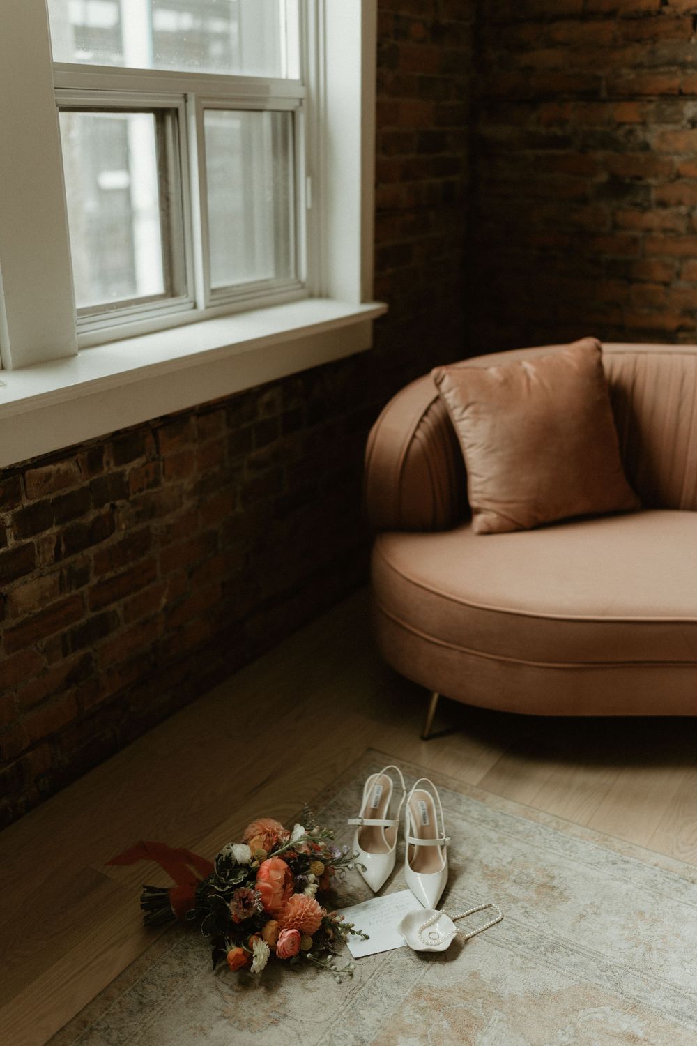 A bridal bouquet of peach roses and white heels rests on the floor beside a soft pink couch near a sunny window.
