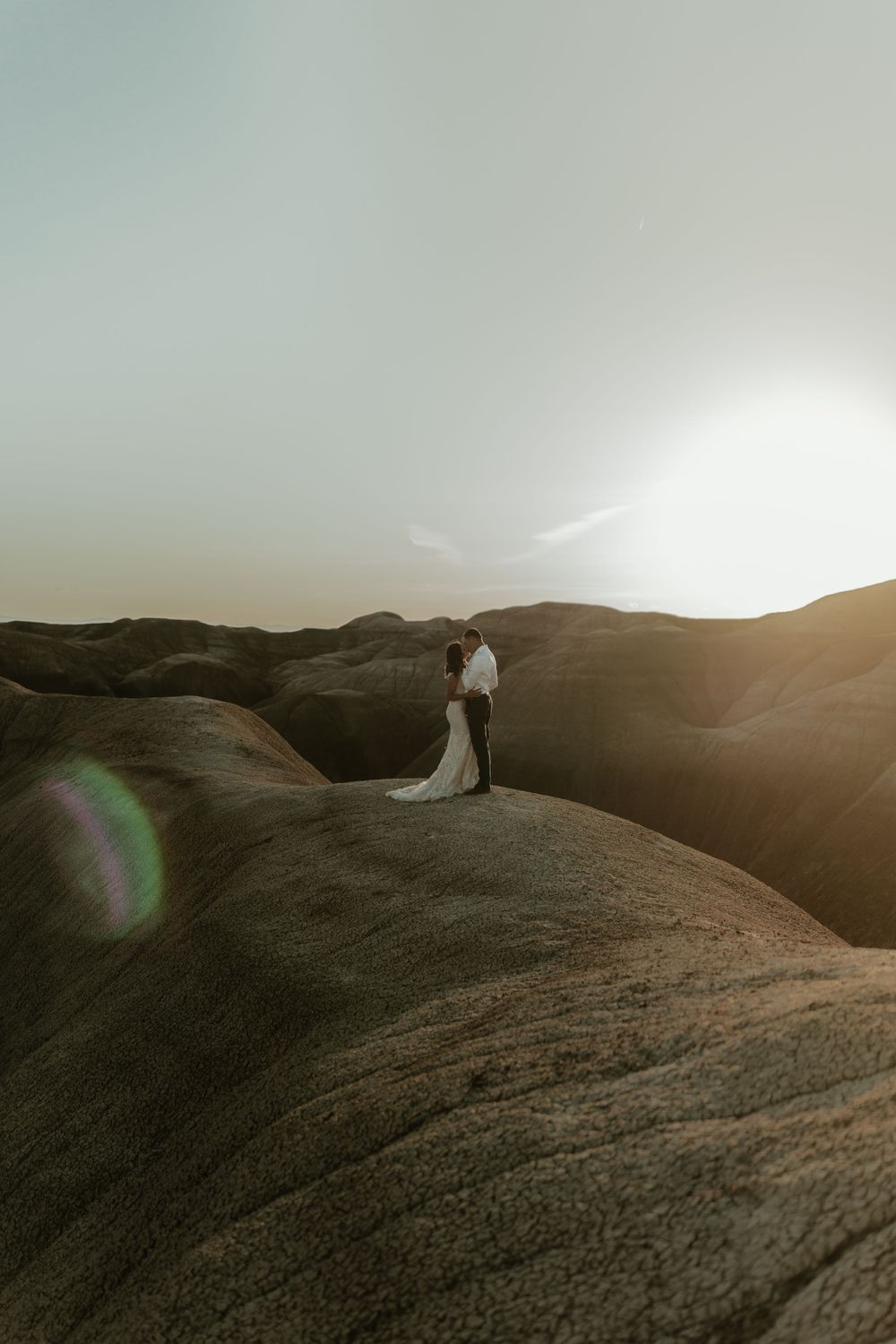 A couple stands silhouetted against the sunset on a rugged mountain ridge in an ethereal desert landscape.