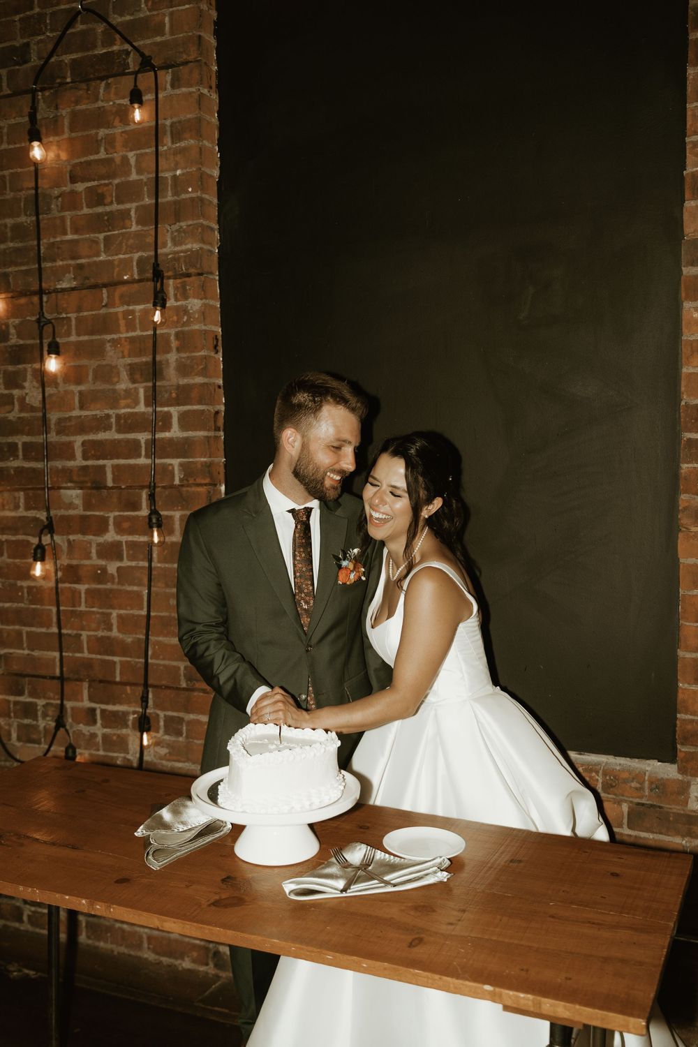 A newly married couple cuts the wedding cake at a rustic wooden table with exposed brick walls and pendant lighting in the background.