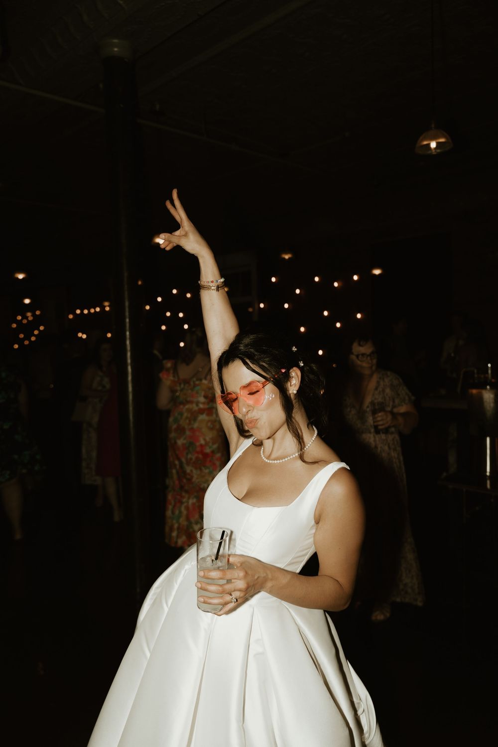A bride in a white wedding dress dances at a reception while holding a drink with their arm raised in celebration.