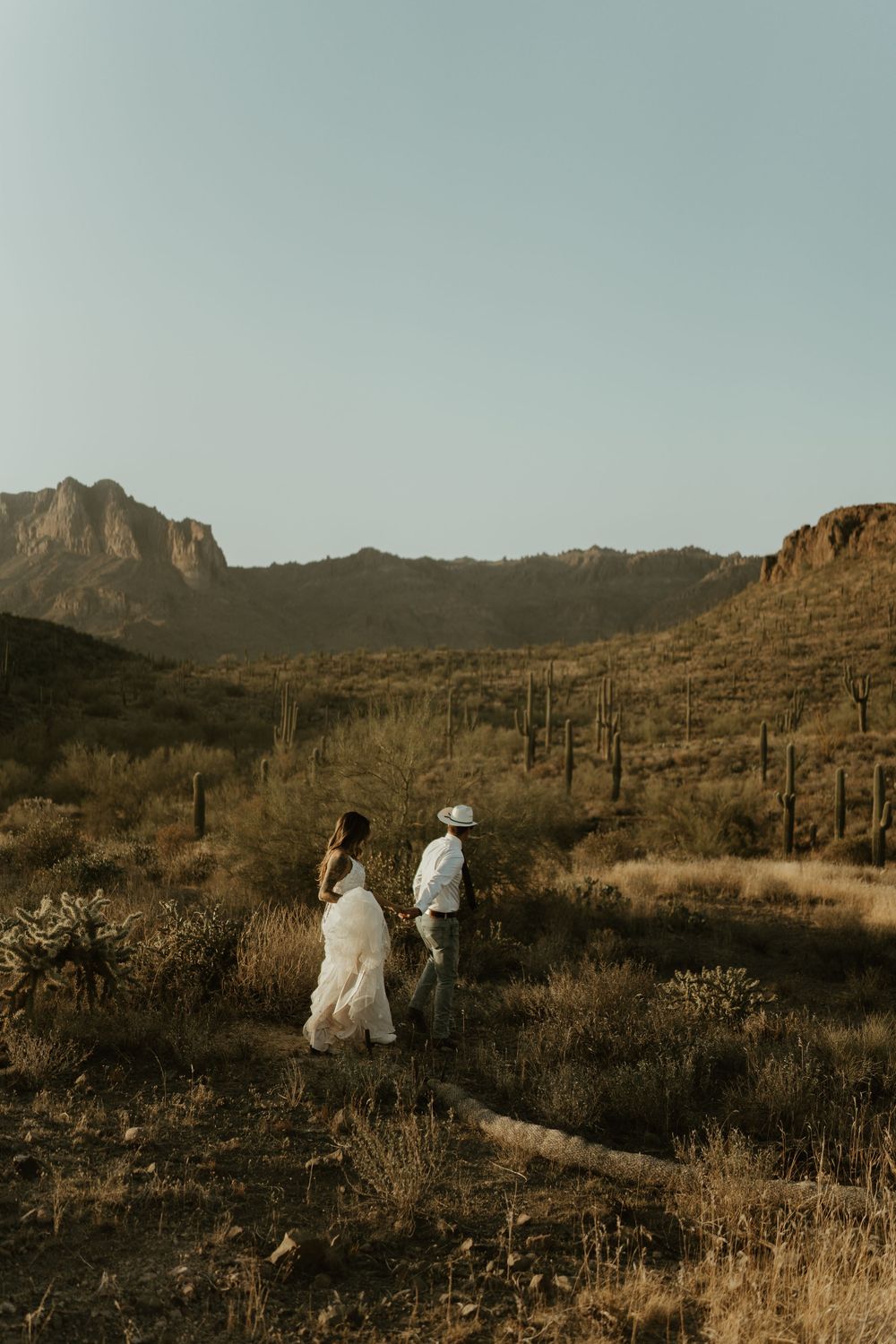 Two figures stand together in a desert landscape with rugged mountains and saguaro cacti at golden hour.