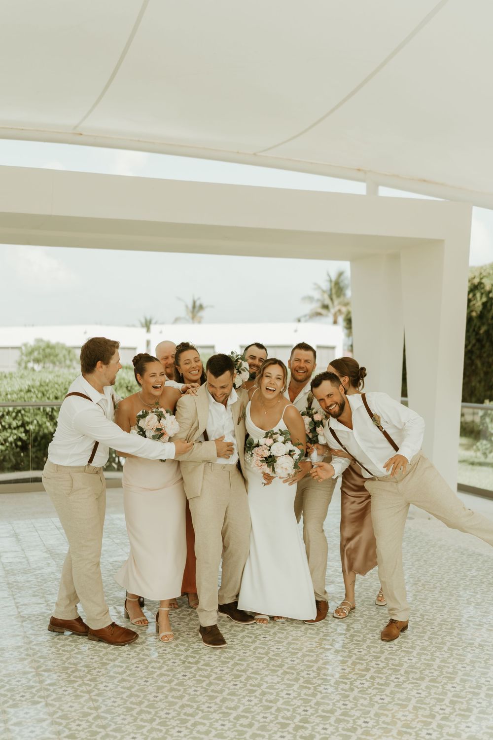 Wedding party in light beige and white attire laughs together while celebrating outdoors at a tropical venue.