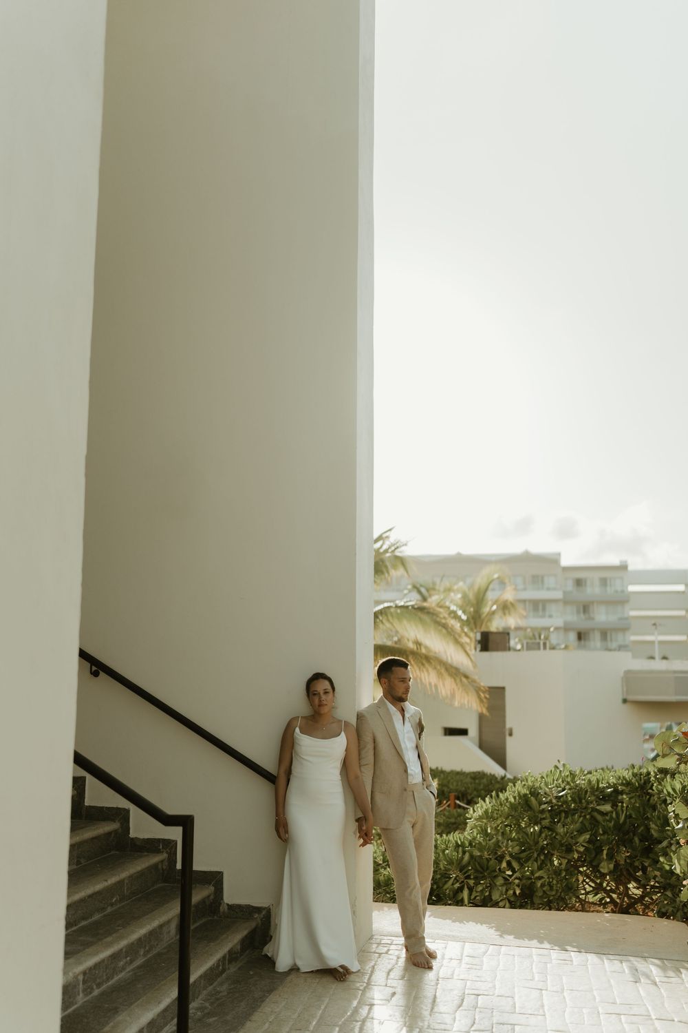 A newlywed couple stands together at an outdoor staircase with palm trees and a modern building in the background.