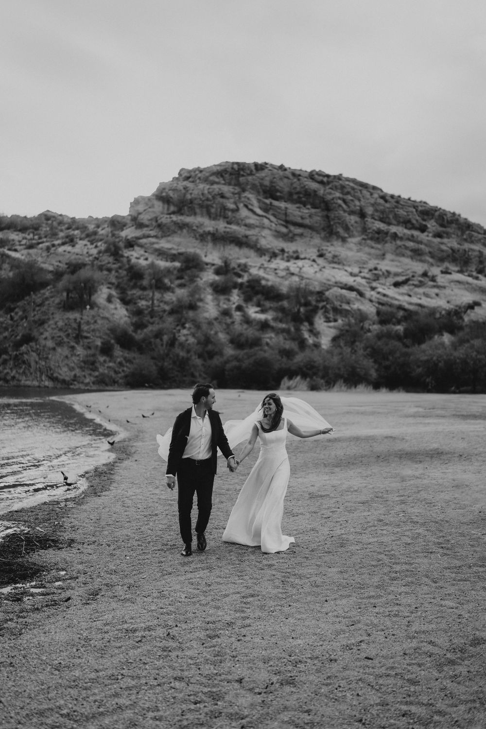 A couple in wedding attire walks hand in hand along a scenic shoreline with rugged cliffs in the background.