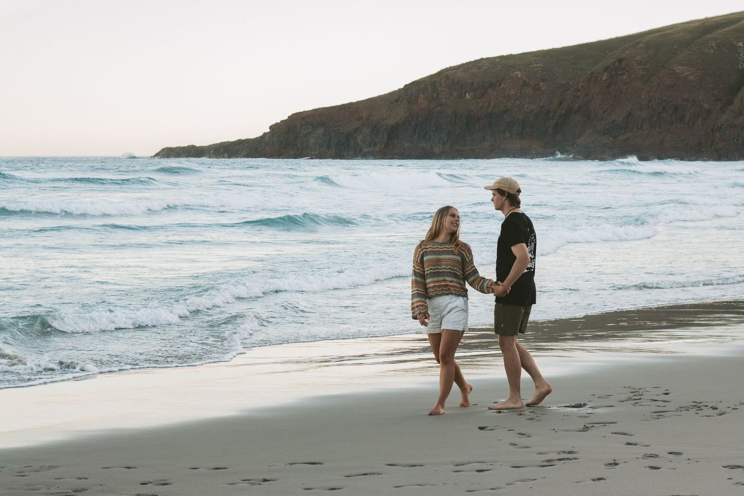 Two people walking barefoot along a scenic beach with cliffs in the background.
