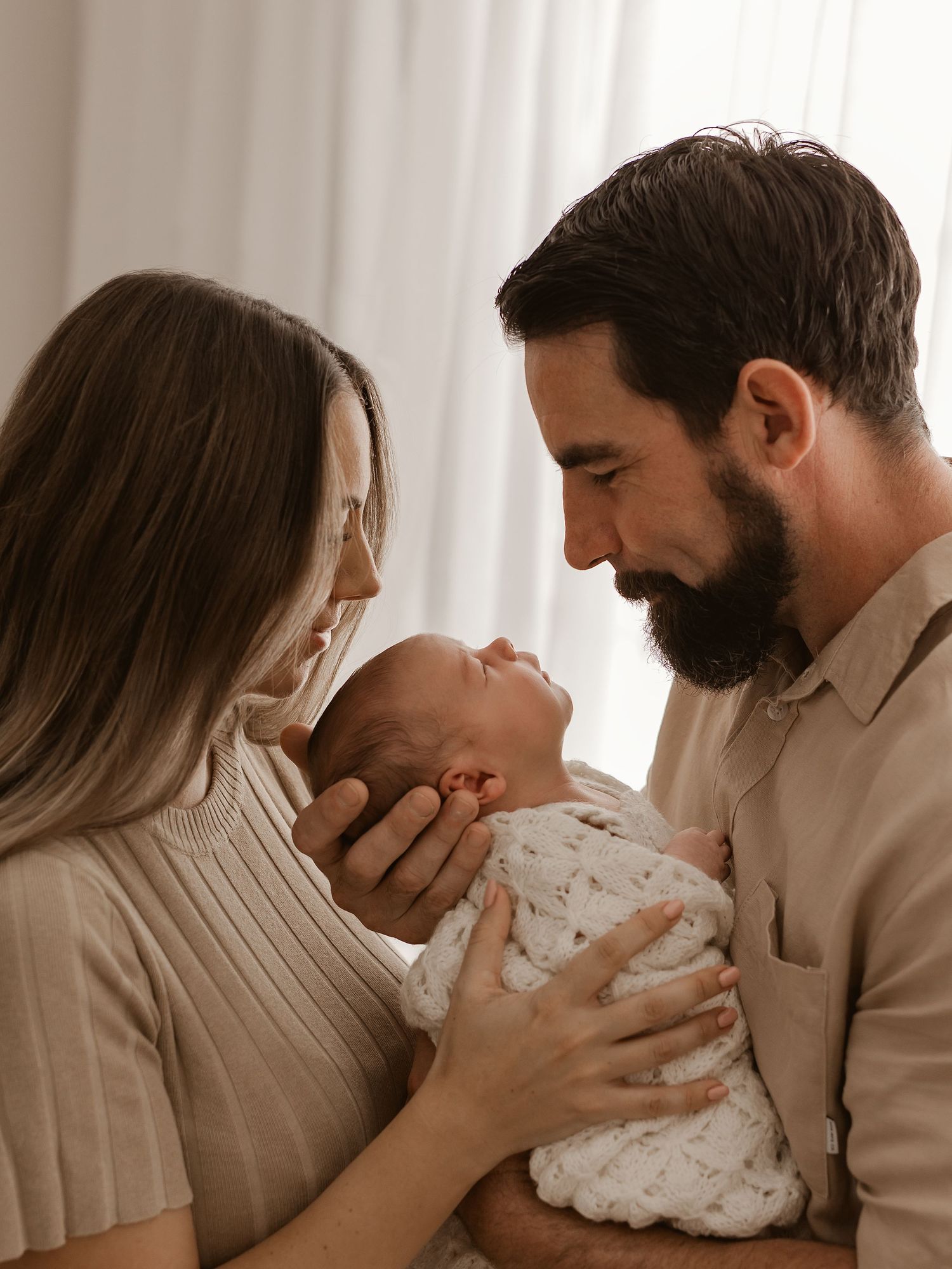 Parents sharing an intimate bonding moment with their newborn baby in warm natural lighting by a window.