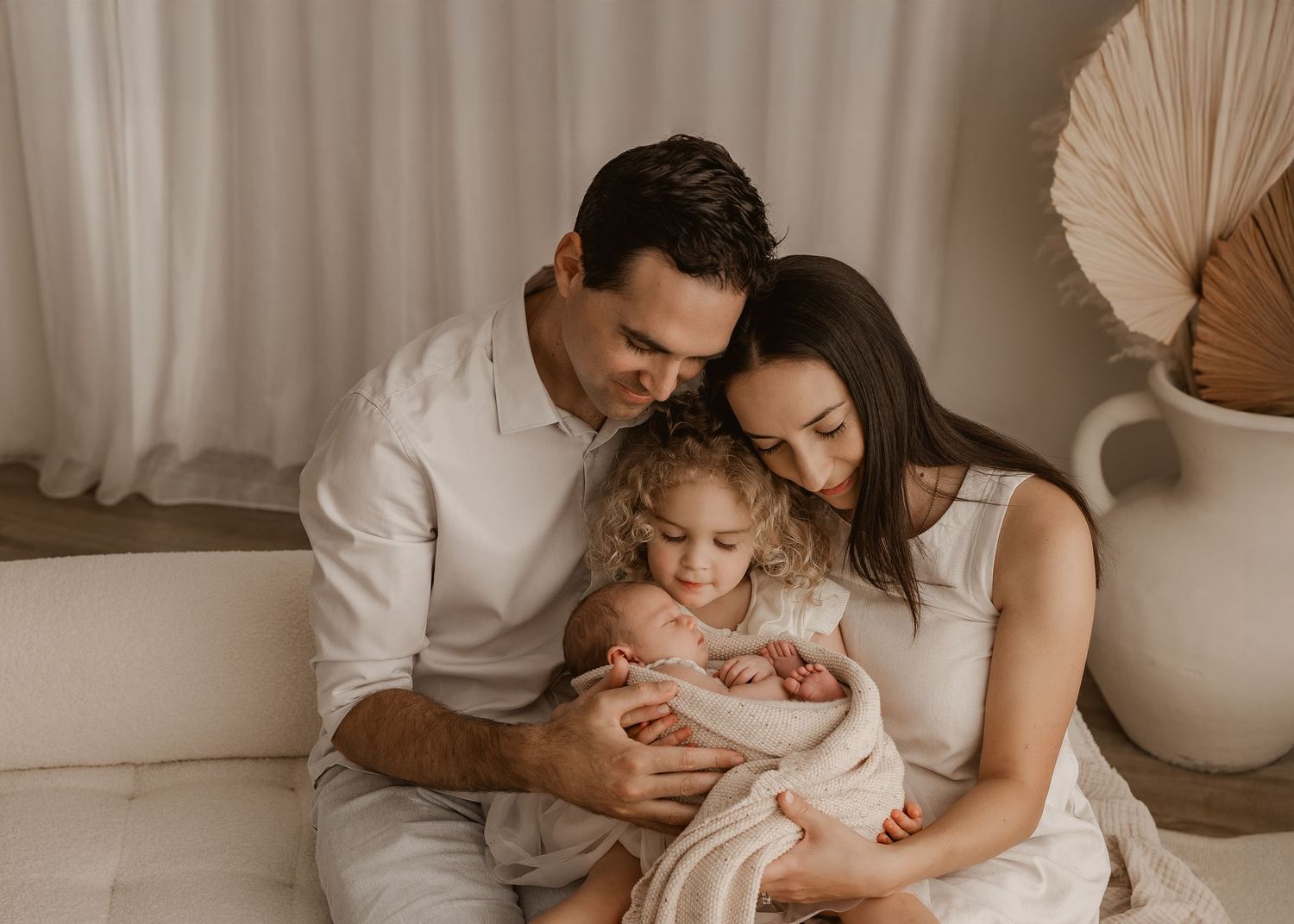 A family sitting together on a cream colored couch in a softly lit room with bohemian decor and neutral tones.