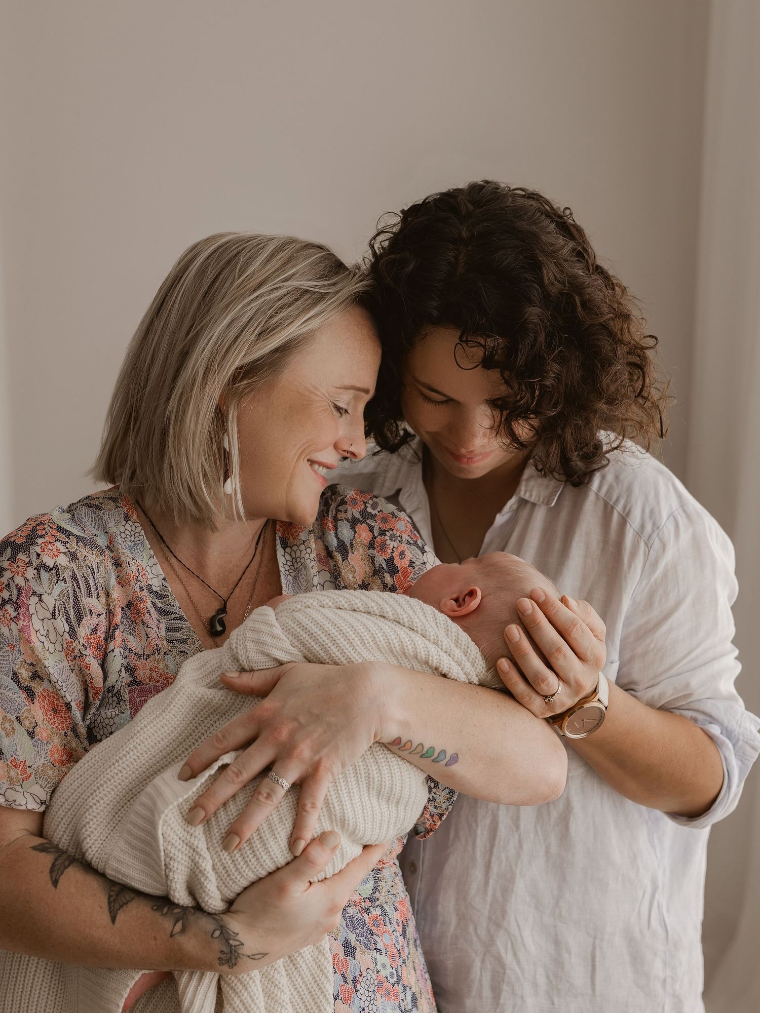 Two people share a tender moment while holding a swaddled newborn baby against a neutral wall background.