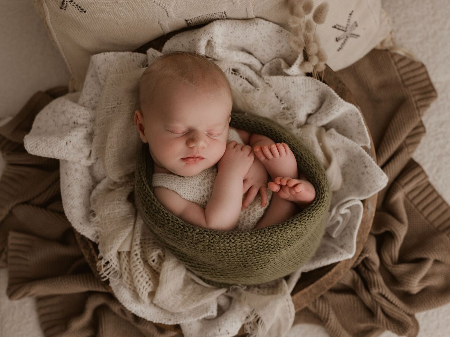 A sleeping newborn swaddled in olive green fabric lying on neutral beige blankets in professional portrait setting.