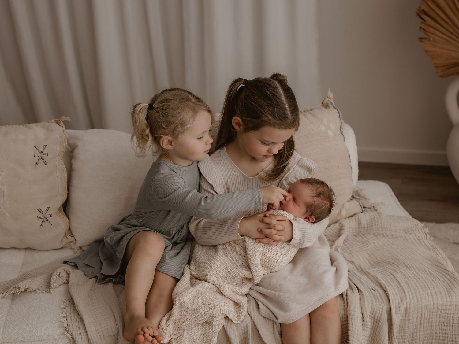 Three children sit together on a cream colored couch, with the older ones tenderly cradling a newborn baby.